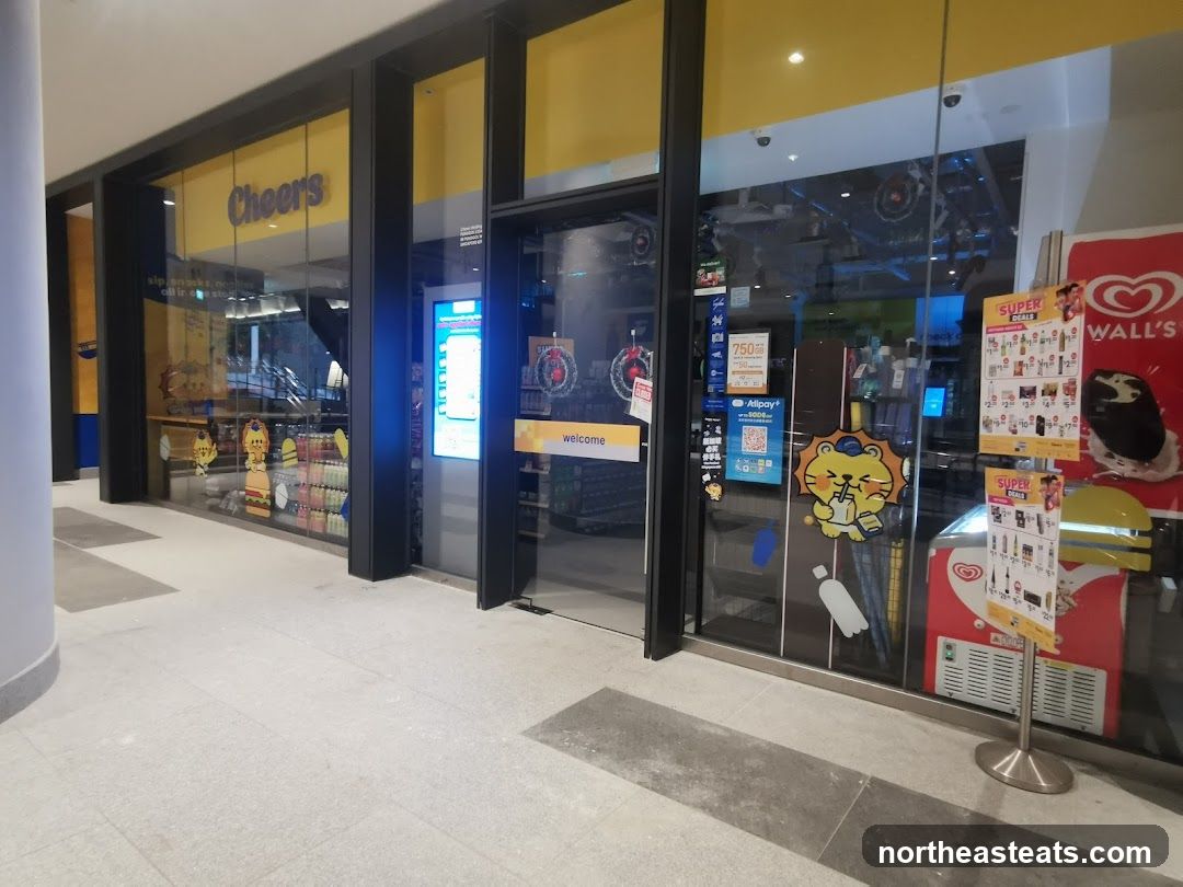 The exterior of a Cheers convenience store with large glass windows and a yellow and blue color scheme. The store's name, "Cheers," is visible above the entrance. Inside, shelves stocked with various products are visible. Promotional posters and a Wall's ice cream freezer are displayed outside the store.