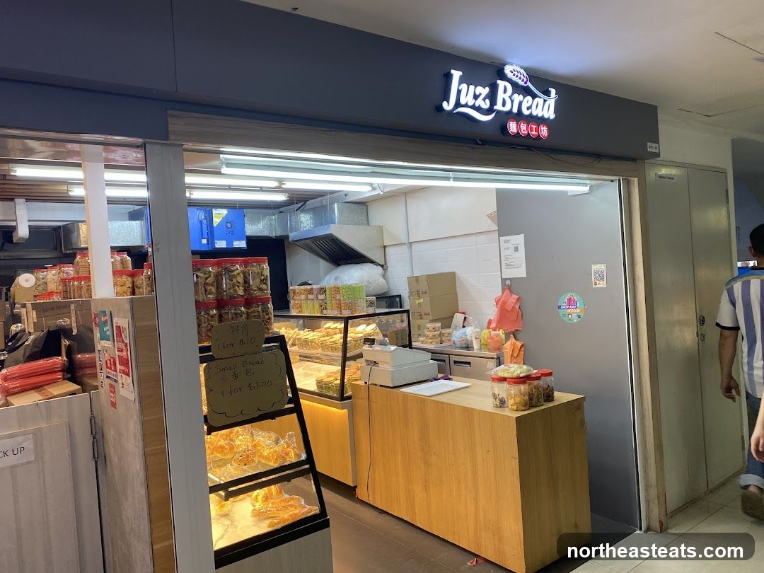 The exterior of a bakery with a prominent sign that reads "Juz Bread" and Chinese characters below. Inside, baked goods are displayed in glass cases and on shelves. There are also jars of snacks and a wooden counter with a cash register. A sign indicates "Pick Up" and another sign lists prices for items like "Special Offer 1 for $10" and "Small Bread 1 for $1.00".