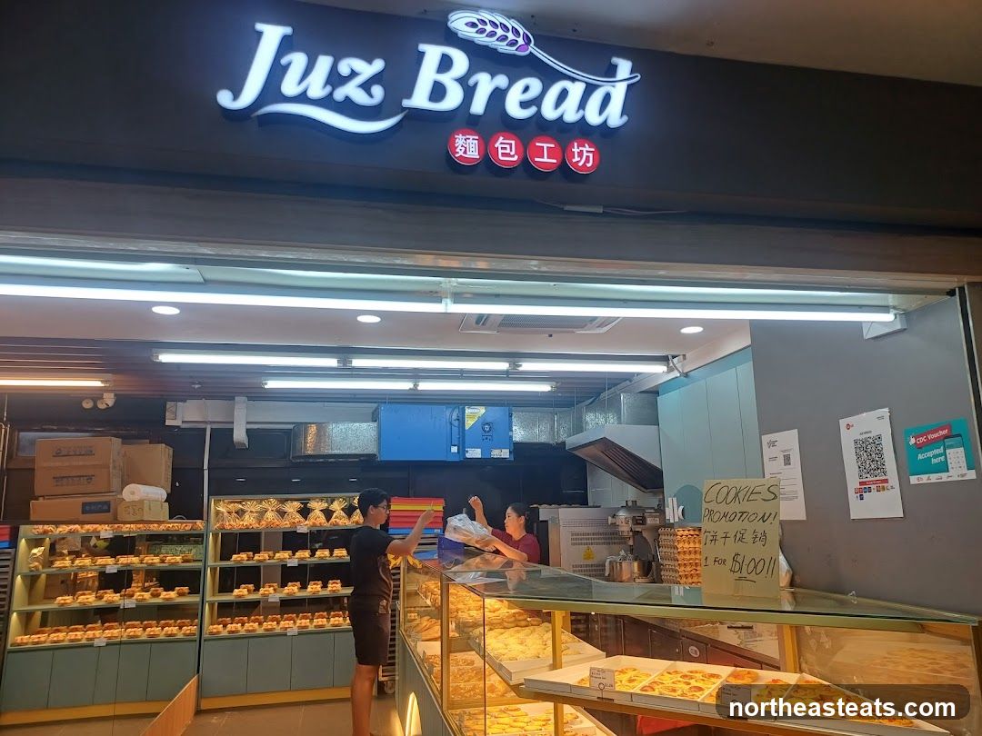 The exterior of a bakery with a prominent "Juz Bread" sign. Inside, shelves are stocked with various baked goods, and two staff members are visible. A sign advertises a "COOKIES PROMOTION! 1 FOR $1.00".