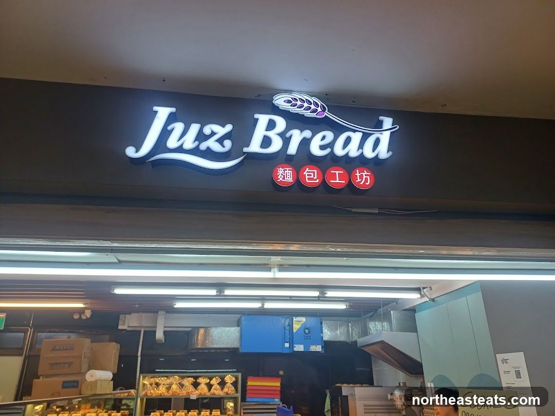 A brightly lit storefront with a prominent illuminated sign that reads "Juz Bread" in English, accompanied by Chinese characters below. The sign features a stylized wheat stalk graphic. Below the sign, the interior of the bakery is visible, showcasing shelves stocked with baked goods and a counter area.