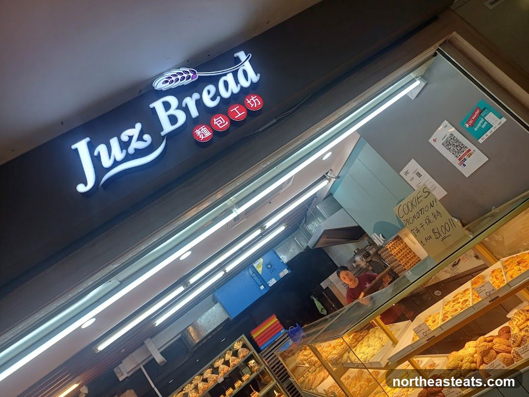 A brightly lit bakery storefront with a prominent "Juz Bread" sign. Inside, a person is visible behind a counter filled with various baked goods displayed in glass cases. A sign advertises a "Cookies Promotion."