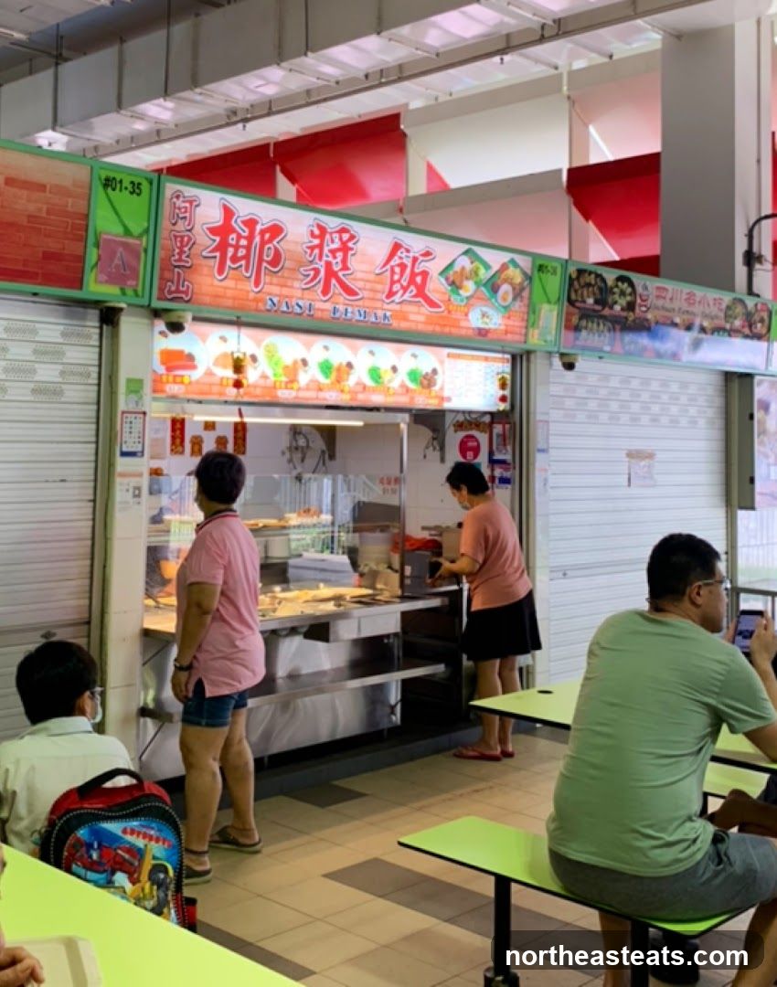 A food stall with a prominent sign displaying "NASI LEMAK" in both Chinese and English. The stall features a display of various food items and customers ordering. The stall is located in an open-air food court setting with tables and chairs.