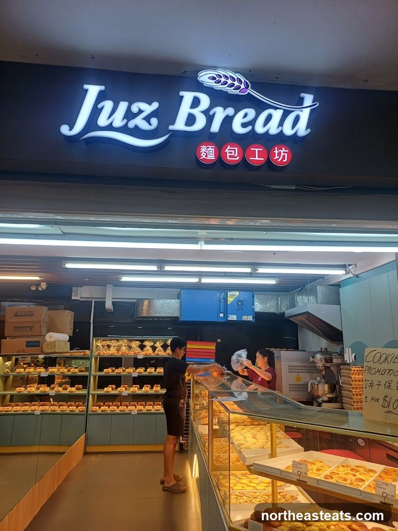 A well-lit bakery storefront with a prominent illuminated sign that reads "Juz Bread" in English and Chinese characters. Inside, shelves are stocked with various baked goods, and two staff members are visible behind a display counter.
