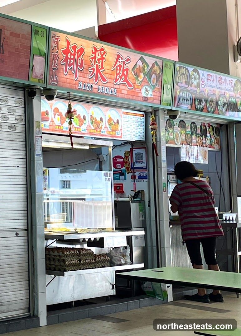 A food stall with a prominent sign displaying "NASI LEMAK" in both Chinese and English. Below the main sign, there are images of various dishes with prices. To the right, another section displays images of Sichuan Famous Delights. A person is standing at the counter, facing away from the camera. The stall has metal shutters on the left side and a display counter with food items and stacks of egg cartons.