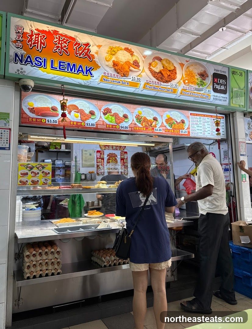A food stall with a large illuminated sign displaying various Nasi Lemak sets and other dishes. The sign features images of the food items with their prices. Below the main sign, there are more food options displayed with prices. Customers are seen ordering at the counter, and staff are preparing food behind the counter. Stacks of eggs are visible on shelves in front of the stall.