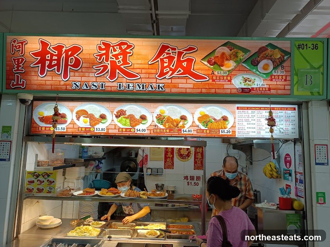 A food stall with a large illuminated sign displaying "NASI LEMAK" in English and Chinese characters. Below the main sign are images and prices for various Nasi Lemak sets and other dishes. A menu board on the right lists prices for Economic Bee Hoo and Nasi Lemak items. Two staff members are visible behind the counter, and a customer is ordering.
