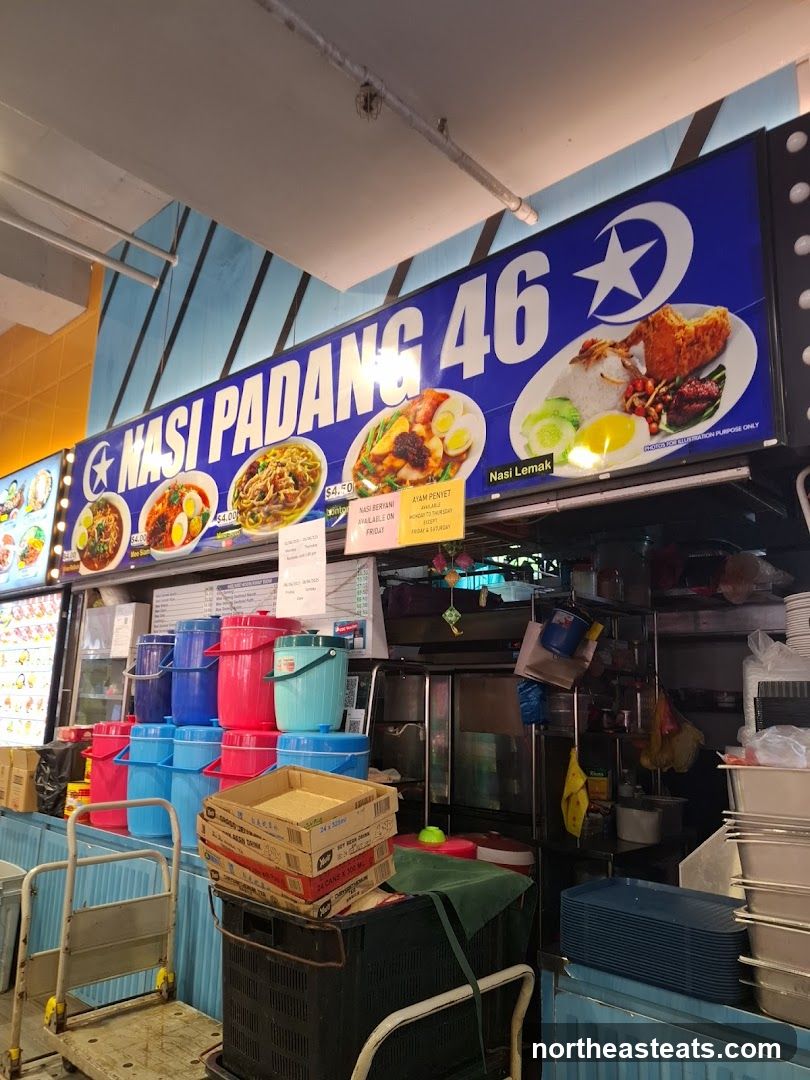 A blue sign with white text and images of food displays the name "NASI PADANG 46". Below the sign, various food items are displayed, and stacks of colorful buckets are visible. The overall scene appears to be a food stall or eatery.