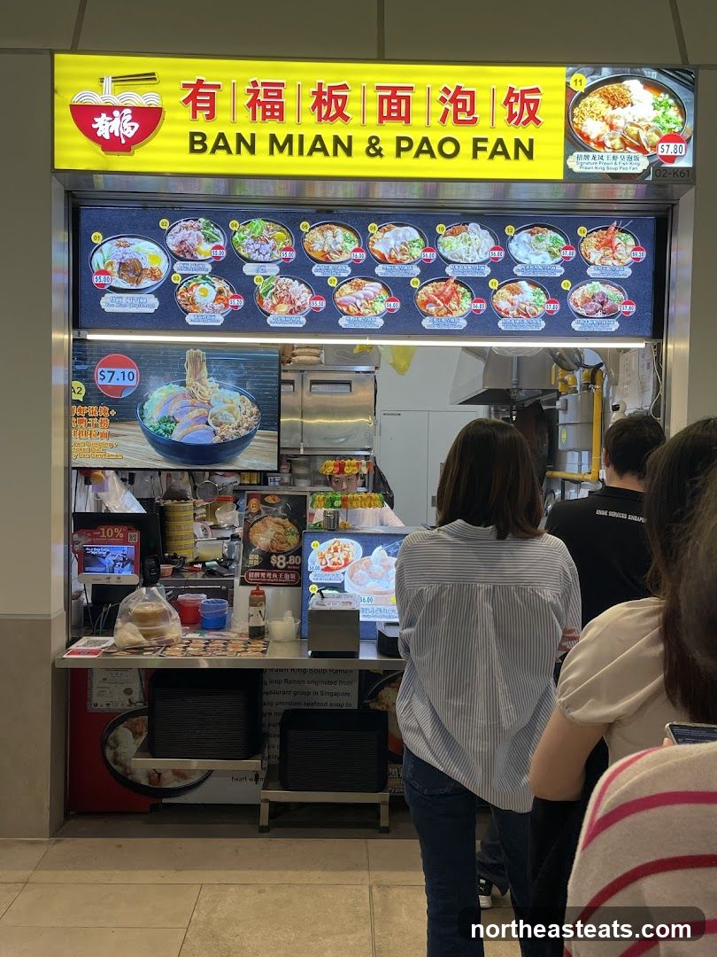 A food stall with a bright yellow sign displaying the name "BAN MIAN & PAO FAN" in English and Chinese characters. Below the sign is a menu board with pictures and prices of various noodle and rice dishes. A large image of a noodle soup bowl is displayed prominently on the left, with a price of $7.10. Other menu items are priced between $5.00 and $9.80. A few customers are visible queuing at the stall.