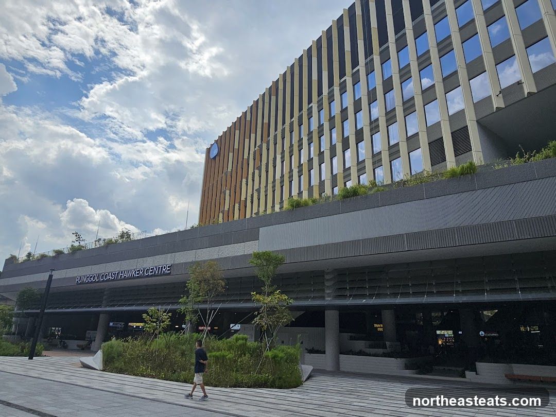A wide shot of the Punggol Coast Hawker Centre building on a cloudy day. The building has a modern design with a facade of vertical wooden slats and reflective windows. A sign above the entrance reads "PUNGGOL COAST HAWKER CENTRE". A person walks on the paved walkway in front of the building.