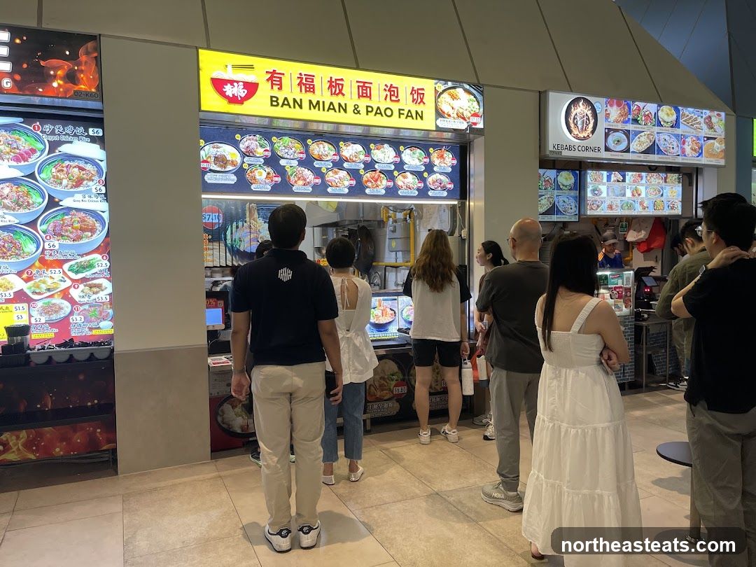 A food stall with a bright yellow sign displaying "BAN MIAN & PAO FAN" in English and Chinese characters. Below the sign, a menu board shows various dishes with pictures and prices. Several customers are standing in line, waiting to order. To the right, another stall labeled "KEBABS CORNER" is visible with its own menu.