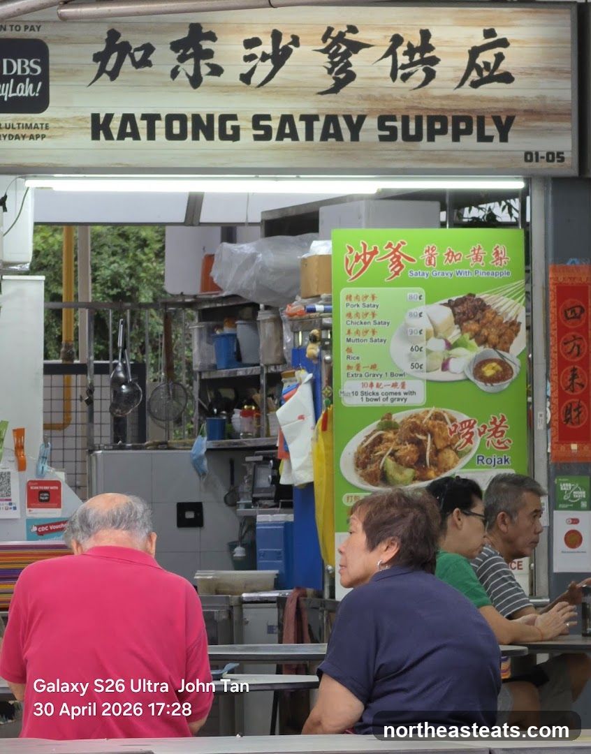 A food stall with a sign that reads "KATONG SATAY SUPPLY" in English and Chinese. There are people sitting at tables in front of the stall. A menu board displays satay and rojak options.