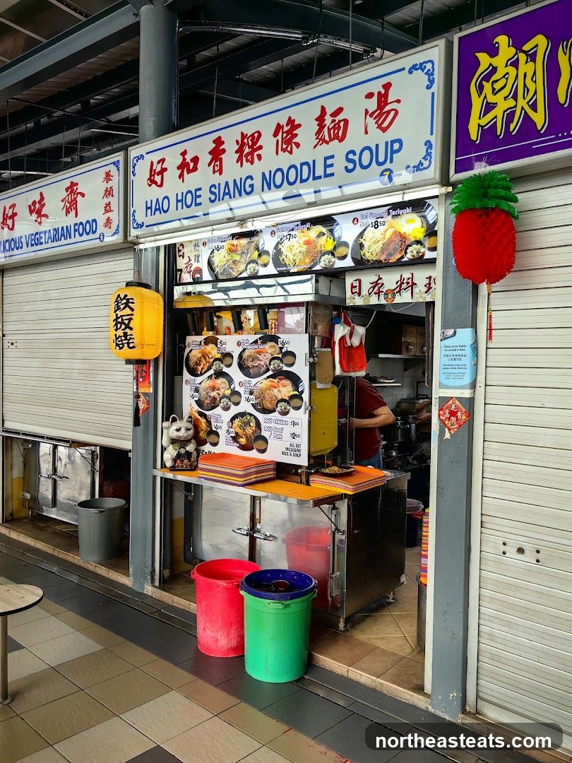 A food stall with a sign that reads "Hao Hoe Siang Noodle Soup" and "Delicious Vegetarian Food". There are pictures of various noodle dishes and teriyaki meals displayed. A person is visible in the background preparing food.