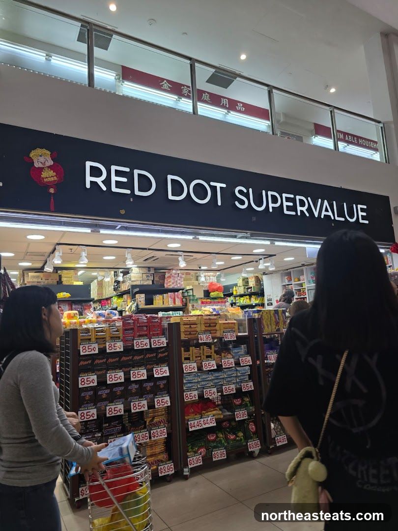 The exterior of a supermarket with a large sign that reads "RED DOT SUPERVALUE". Below the sign, shelves are stocked with various products, including candy bars and snacks, with visible price tags. Two women are seen in the foreground, one pushing a shopping cart.