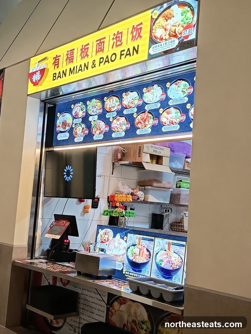 A food stall with a bright yellow sign displaying the name "BAN MIAN & PAO FAN" in English and Chinese. Below the sign is a menu board with pictures and prices of various noodle and rice dishes. The stall has a counter with a cash register, a digital display, and food items.