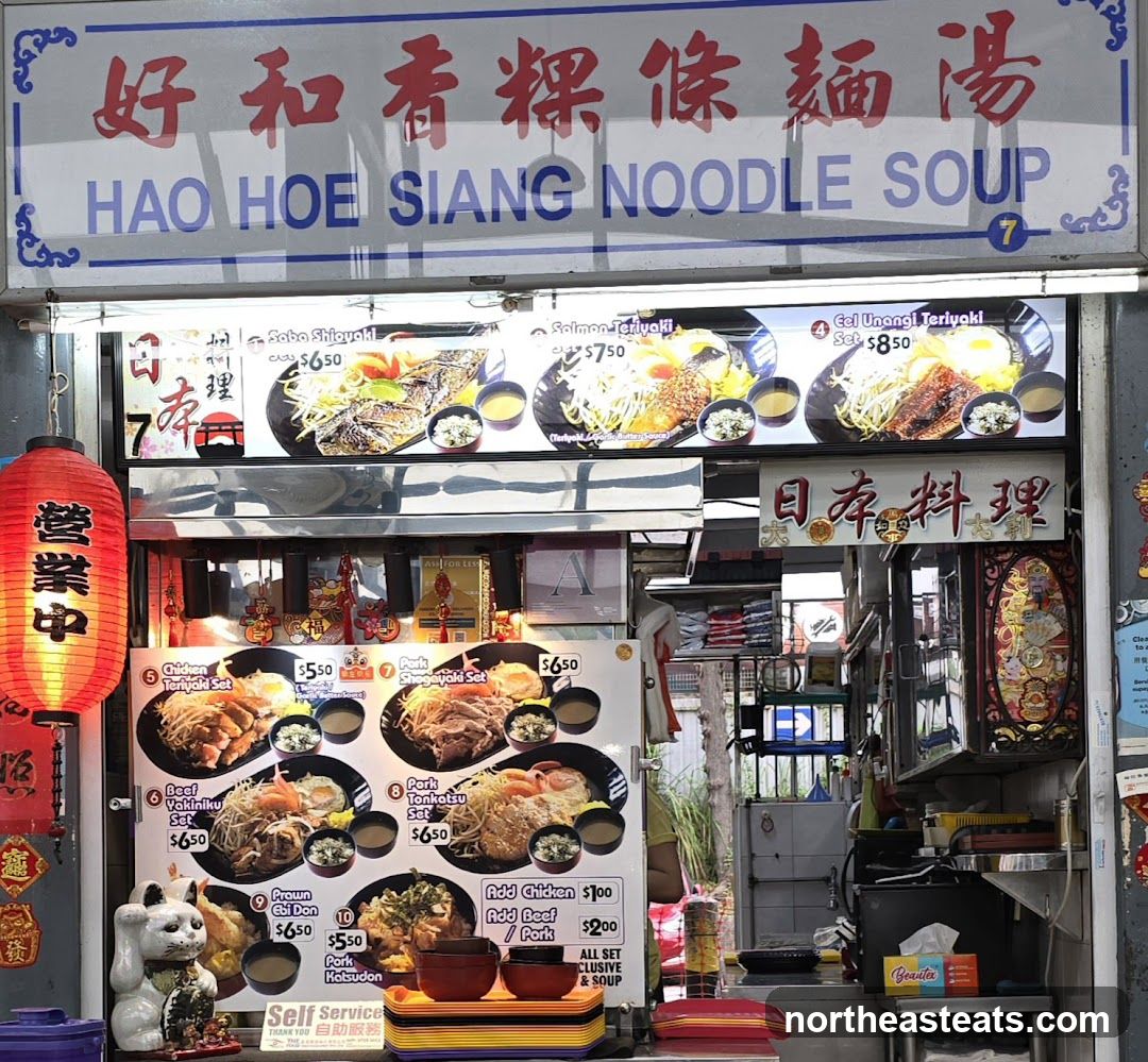 A food stall with a large sign displaying the business name and menu items. The stall features various dishes with prices, including teriyaki sets, yakiniku, and donburi. A red lantern hangs to the left, and a lucky cat figurine is placed near the bottom.