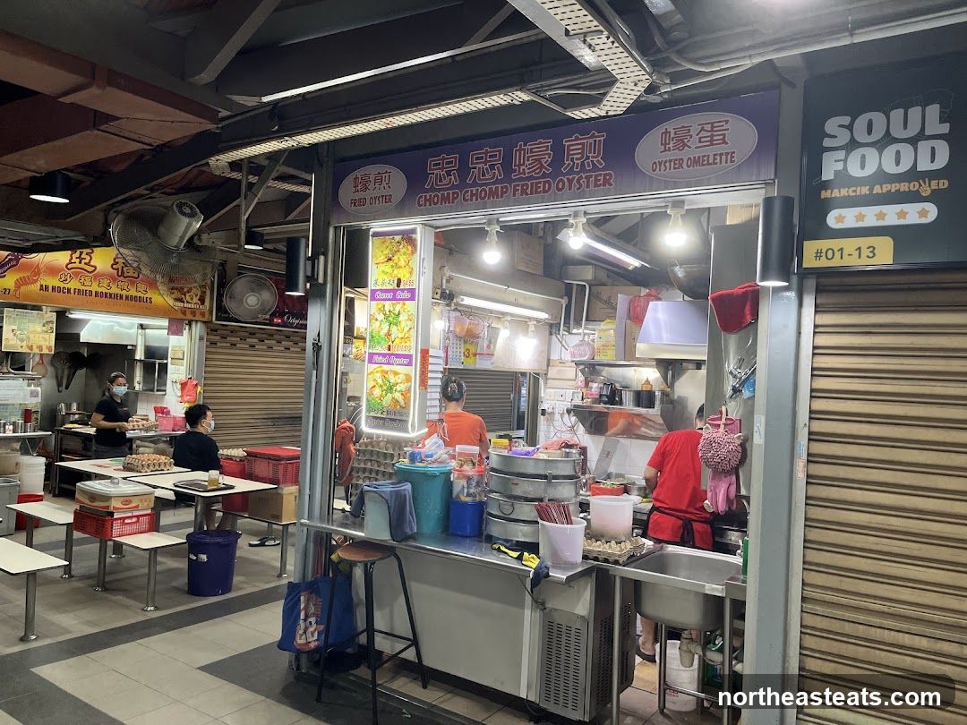 A food stall with a sign that reads "Chomp Chomp Fried Oyster" and "Oyster Omelette". There are people working behind the counter and customers seated at tables. The stall is part of a larger food court or market.