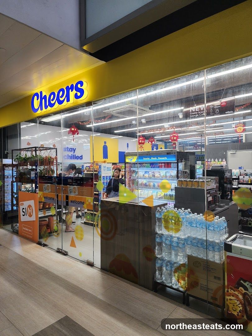 The exterior of a Cheers convenience store with a bright yellow facade and a large blue "Cheers" sign. The entrance has glass doors with "welcome" signs and "SIM" branding. Inside, shelves are stocked with various products, and a staff member is visible behind the counter.