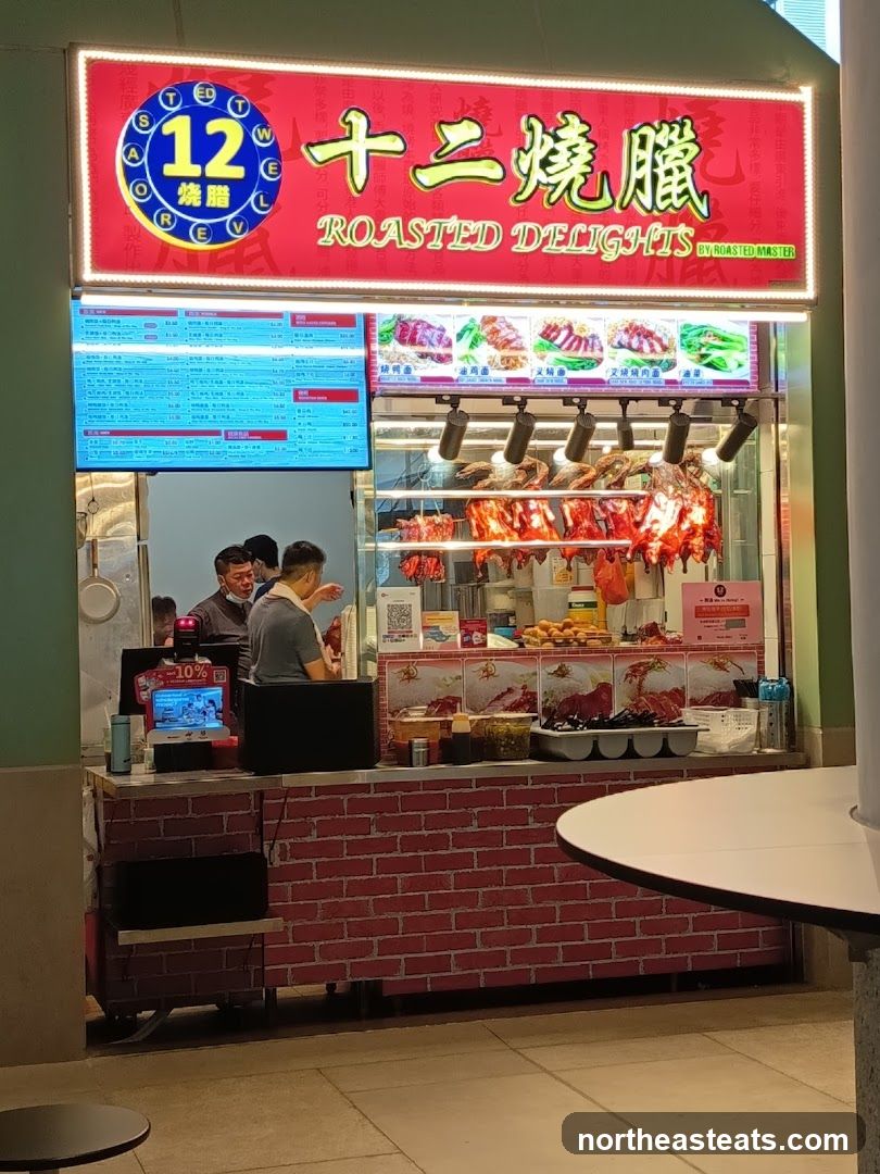A food stall with a bright red sign displaying Chinese characters and the English text "ROASTED DELIGHTS BY ROASTED MASTER". Below the sign are images of roasted meats and a digital menu board. The stall displays various roasted ducks and other meats hanging behind a counter.