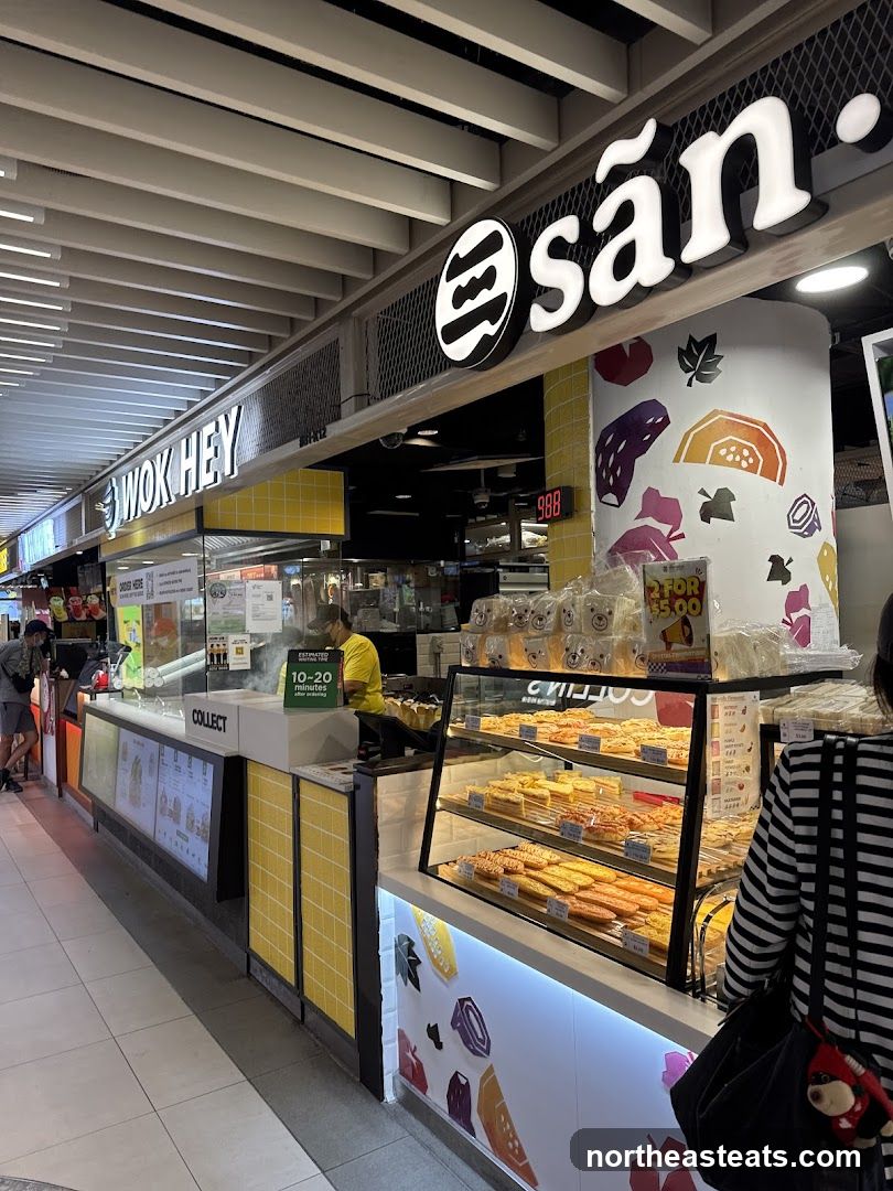 A view of two adjacent food stalls, one labeled "WOK HEY" and the other "sān". The "WOK HEY" stall features a yellow tiled counter with a "COLLECT" sign and a digital display showing an estimated waiting time of 10-20 minutes. The "sān" stall has a display case filled with pastries and baked goods. Both stalls have prominent signage on the overhead structure.