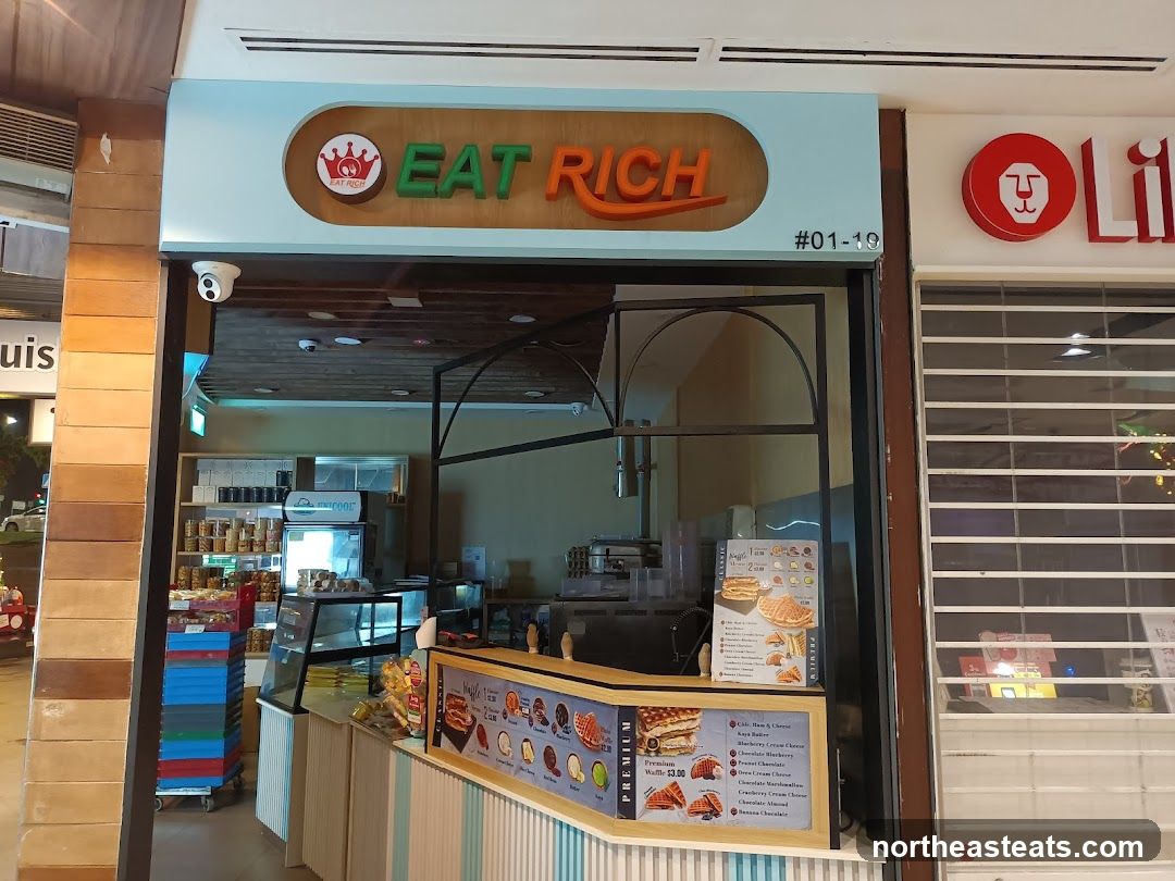 The storefront of a food establishment named Eat Rich. The shop has a light blue facade with the name "EAT RICH" in orange and green letters above the entrance. A menu board is visible inside the shop, displaying various waffle options and prices.