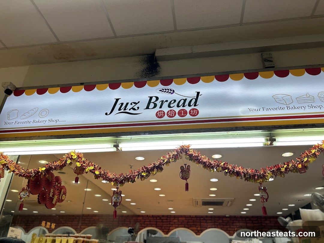 A brightly lit bakery storefront with a prominent sign that reads "Juz Bread" and "Your Favorite Bakery Shop". The sign features illustrations of bread, cakes, and pastries. Below the sign, festive red and gold garlands with small mouse ornaments and red lanterns are hung, suggesting a holiday or celebratory season.