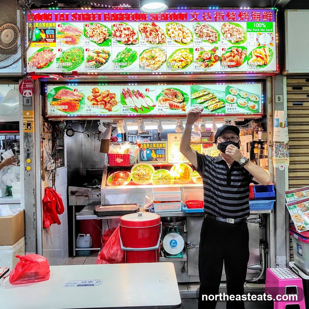 A man stands in front of a food stall with a large illuminated menu board displaying various seafood dishes. The menu board features images and prices of items like Stingray, Prawn Roll, Tiger Prawn, and Garlic Scallops. The stall also advertises "100% No Pork No Lard" and "Since 1985".