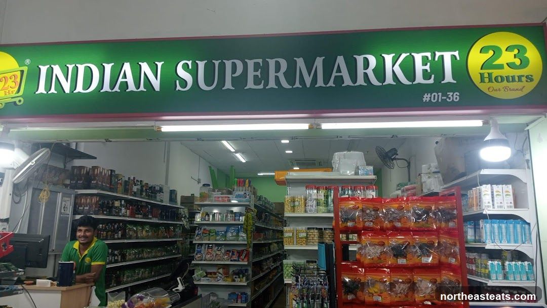 The exterior of an Indian Supermarket with a large green sign above the entrance. The sign reads "INDIAN SUPERMARKET 23 Hours Our Brand" and includes the unit number "#01-36". Inside the supermarket, shelves are stocked with various products, and a male employee is visible behind a counter.