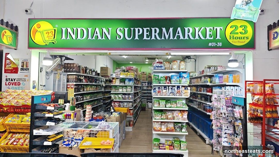 The exterior of an Indian supermarket with its name prominently displayed on a green sign. The store is open, revealing aisles stocked with various products. Baskets of goods are visible near the entrance.