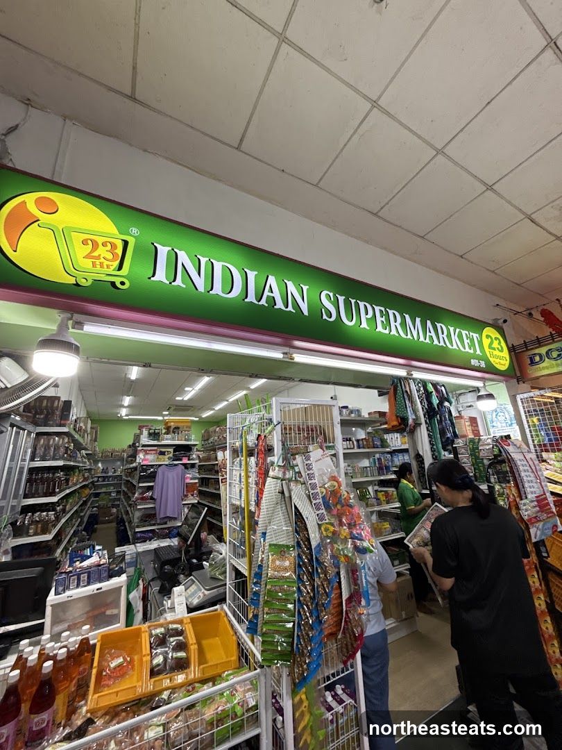 The exterior of an Indian supermarket with a prominent green sign displaying "INDIAN SUPERMARKET" and a "23 Hours" logo. The entrance is open, revealing aisles stocked with various goods, including bottles, packaged items, and shelves filled with products. Two individuals are visible inside the store.