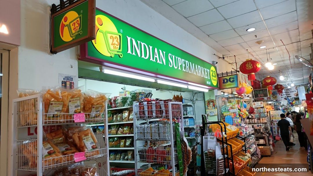 The exterior of an Indian Supermarket with a large green sign that reads "INDIAN SUPERMARKET 23 Hours". There are shelves stocked with various food items and snacks visible through the open storefront. Red lanterns hang from the ceiling.