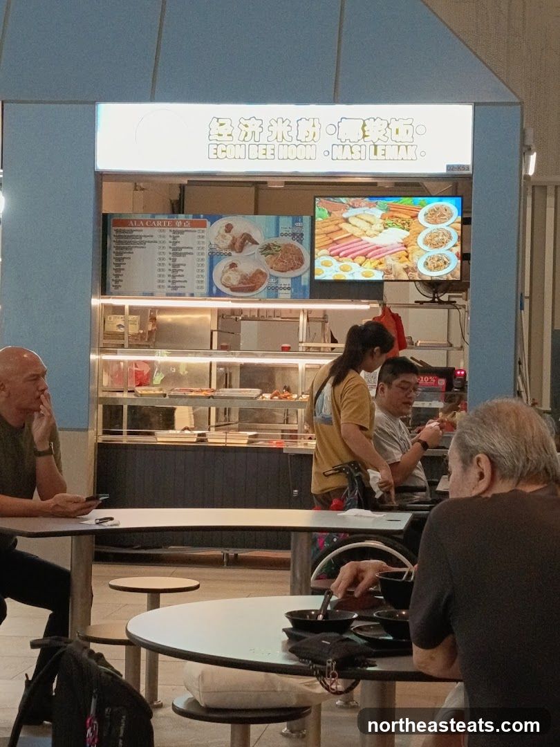 A food stall with a bright sign displaying "ECON BEE HOON - NASI LEMAK" in English and Chinese. Below the sign, there is an "ALA CARTE" menu board and a digital display showing images of food. Several people are seated at tables in front of the stall.