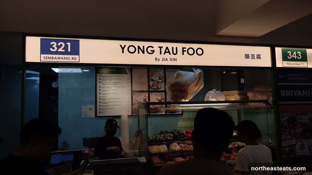 A storefront for a Yong Tau Foo stall. The stall has a large illuminated sign that reads "YONG TAU FOO BY JIA XIN" and also displays the stall number "321 SEMBAWANG RD" and "343 YISHUN AVE 11". Below the main sign, a menu board is visible with various Yong Tau Foo options and prices. The display counter shows fresh ingredients for the dish.