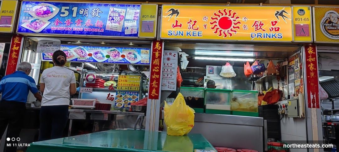 Two food stalls are visible, one on the left specializing in Wanton Egg Noodles and another on the right selling drinks. The stall on the left has a large blue sign with images of noodles and the text "51 明發 全蛋雲吞麵 WANTON EGG NOODLE". A "WE ARE HIRING!!!" sign is also visible. The stall on the right has a yellow sign with "SUN KEE DRINKS" and images of swallows and a sun. A smaller sign lists drinks like "SOYA BEAN", "RED MEST DRINK", and "GRASS JELLY" with prices.