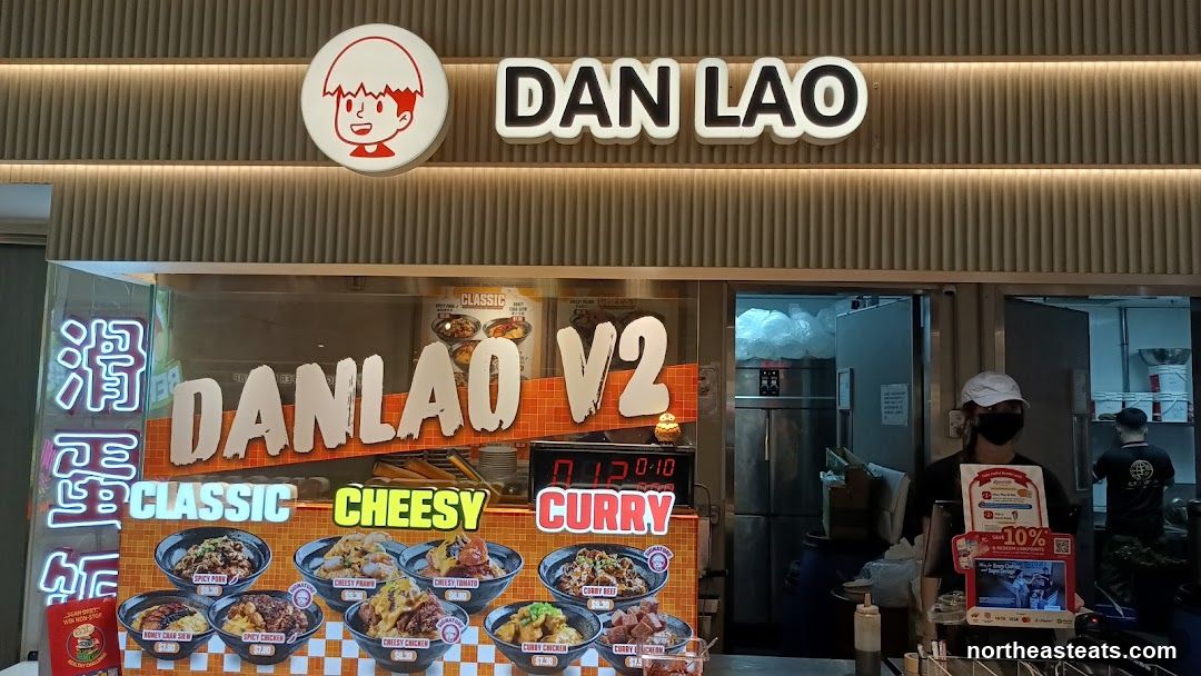The storefront of a restaurant called Dan Lao V2. The sign above the entrance reads "DAN LAO" with a cartoon character logo. Below the sign, a menu display shows various dishes with images and names such as "Spicy Pork", "Cheesy Prawn", "Cheesy Tomato", "Curry Beef", "Honey Char Siew", "Spicy Chicken", "Cheesy Chicken", and "Curry Chicken". A digital timer displays "01:20:10". A staff member wearing a mask and cap is visible behind the counter.
