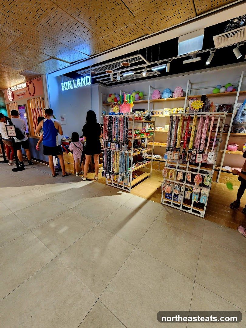 A view of the interior of a store with the sign "FUN LAND" illuminated in neon blue. Shelves are stocked with various items including lanyards, backpacks, and other accessories. Several people are standing in line to the left of the frame, near another establishment with a sign that reads "SWEET CHOON".