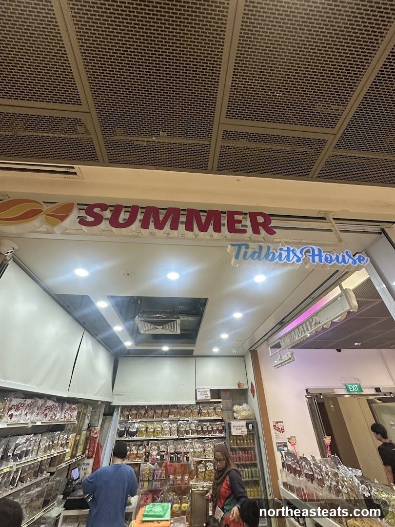 The exterior of a shop called Summer Tidbits House. The shop sells various snacks and tidbits, with shelves stocked full of packaged goods. There are people inside the shop browsing the items.