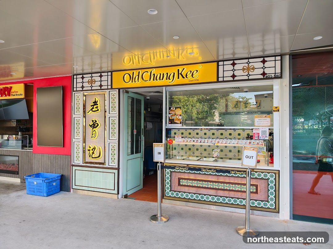 The exterior of an Old Chang Kee shop with a bright yellow sign and traditional Chinese characters. The shop has a glass display counter showing various food items. There are "IN" and "OUT" signs indicating customer flow.