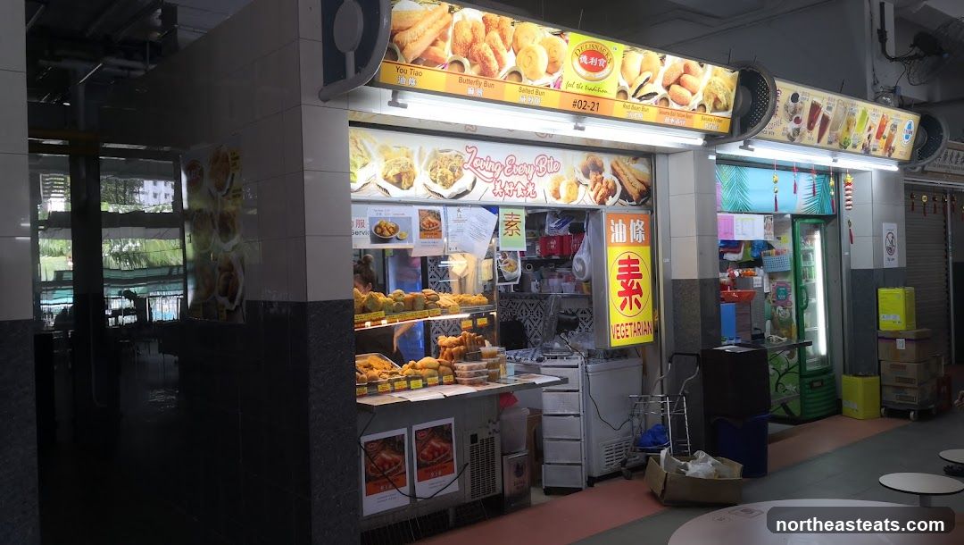A food stall with a variety of fried vegetarian items displayed on shelves and in containers. Above the stall, illuminated signs show pictures of the food items and the stall's name, "DELISNACKS" and "Loving Every Bite". A vertical sign with Chinese characters and the word "VEGETARIAN" is prominently displayed. The stall appears to be part of a larger food court or market.