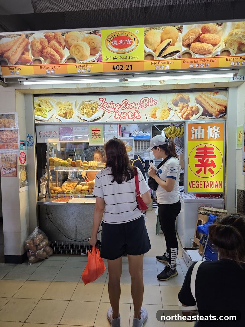 A food stall with a display of various fried snacks and buns. The stall has a prominent sign with the name "DELISNACKS" and images of their offerings. There are also signs indicating "Vegetarian" and "Self Service".