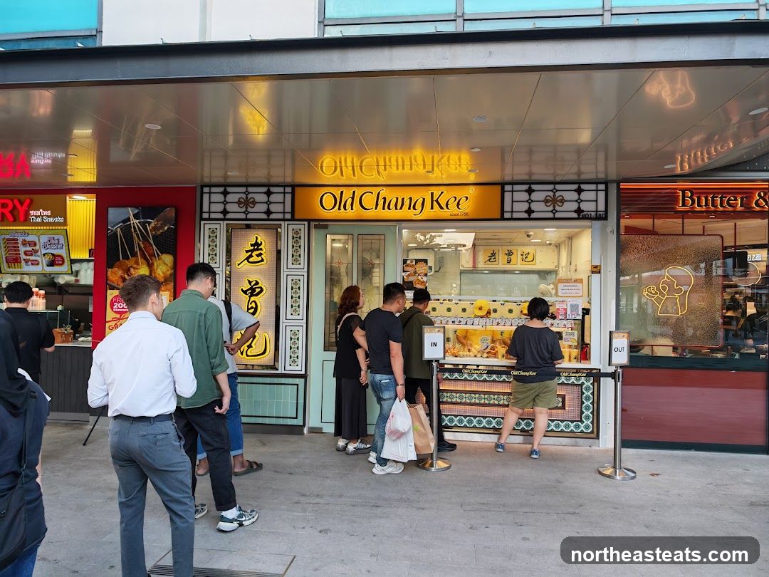A group of people are queuing outside a food stall with a prominent yellow sign that reads "Old Chang Kee". The stall appears to be selling fried snacks.