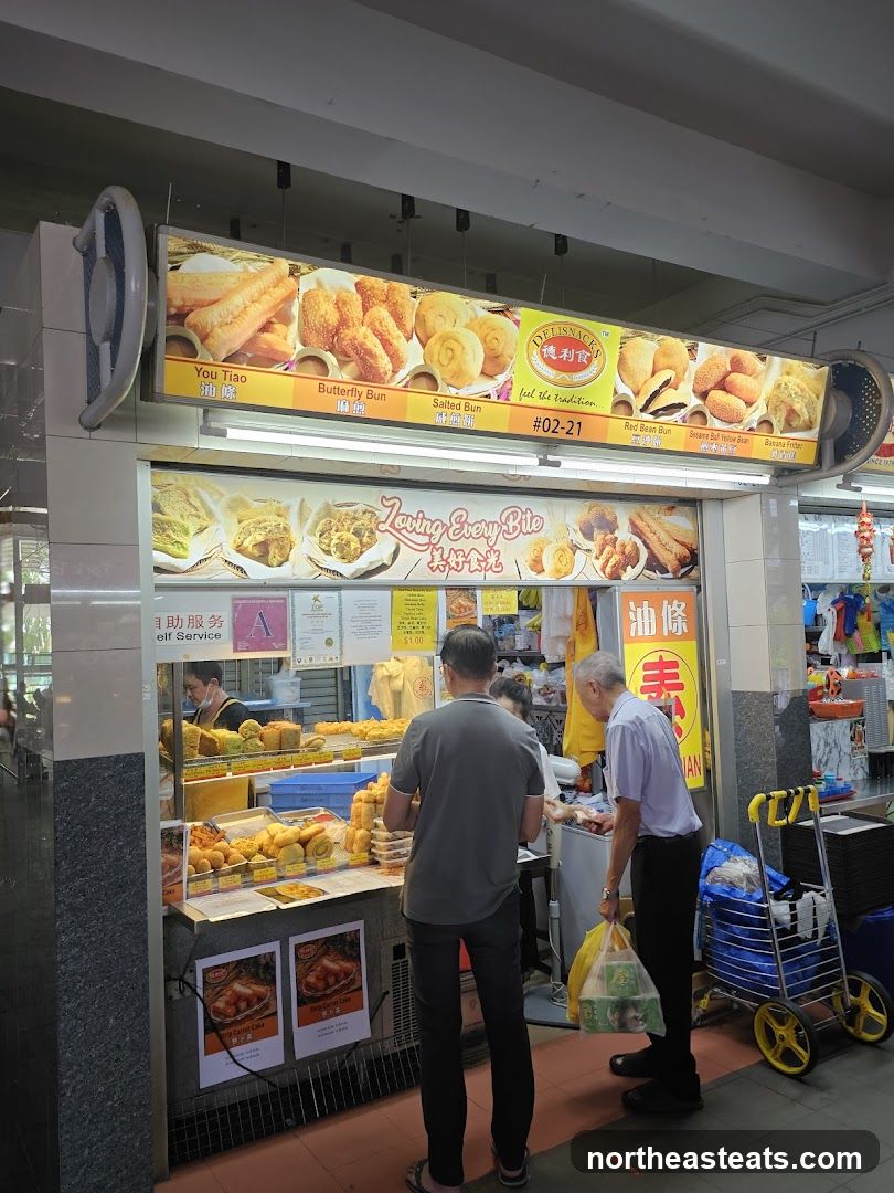 A food stall with a bright overhead sign displaying various fried snacks and buns. The stall has a display counter with a variety of fried food items. Two customers are seen ordering from the stall.
