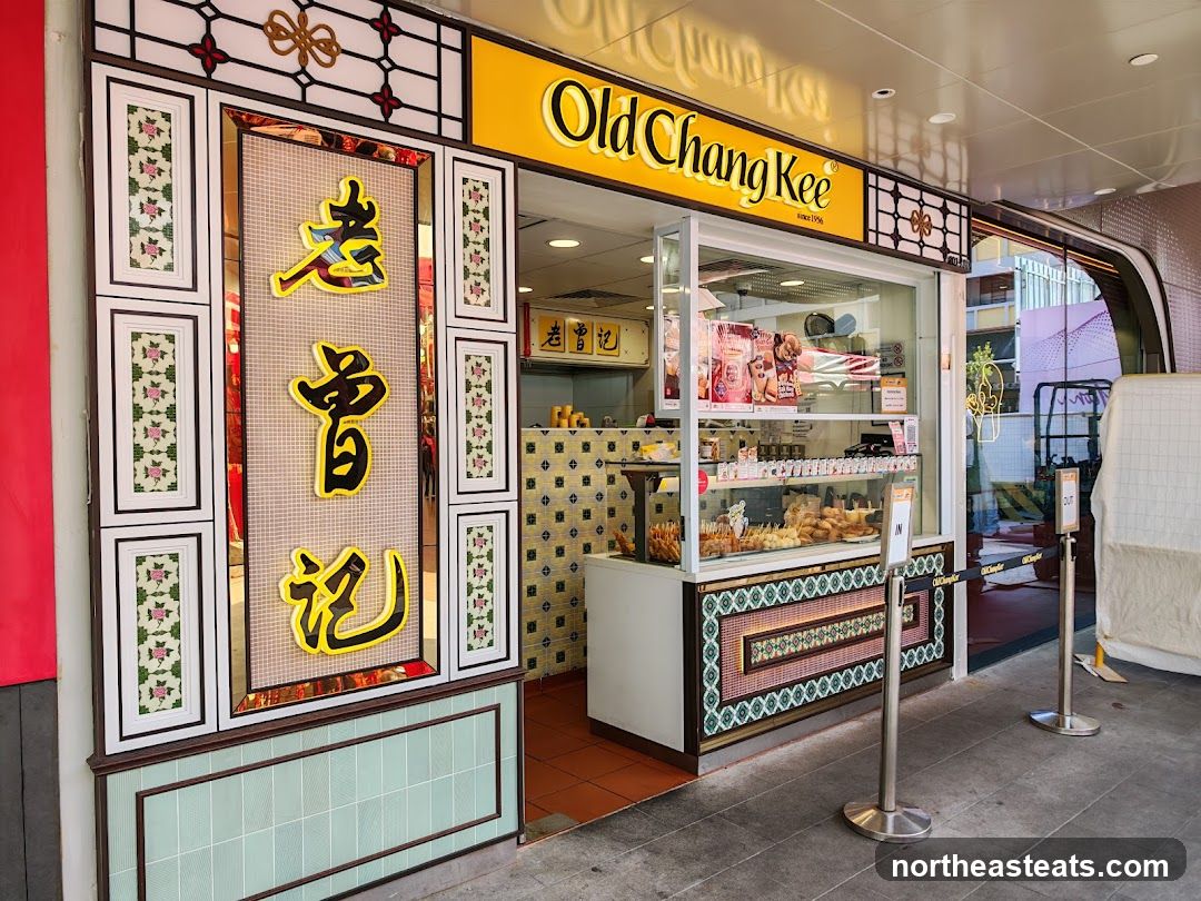 A brightly lit storefront with a yellow awning and the words "Old Chang Kee" in large, illuminated letters. The facade features decorative panels with floral motifs and Chinese characters. A glass display case showcases various food items, and a counter is visible inside. Queue barriers with "IN" and "OUT" signs are placed in front of the entrance.
