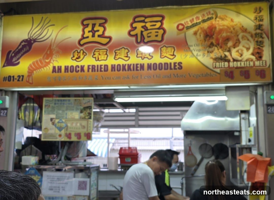 A bright yellow sign with red and yellow Chinese characters and English text advertising "AH HOCK FRIED HOKKIEN NOODLES". The sign features illustrations of a squid and a shrimp, and a picture of the dish. Below the sign, a menu board is visible, along with people working in the food stall. Prices are listed as $4, $5, and $6.