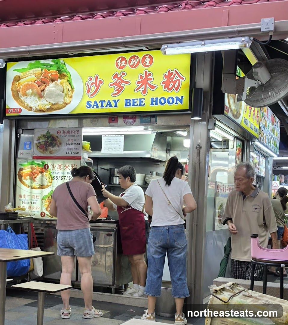A food stall with a large yellow sign displaying "SATAY BEE HOON" in English and Chinese, along with images of the dish. Below the main sign, there are smaller signs with menu items and prices. Several people are seen ordering or waiting for food. The stall appears to be part of an outdoor food court or hawker center.