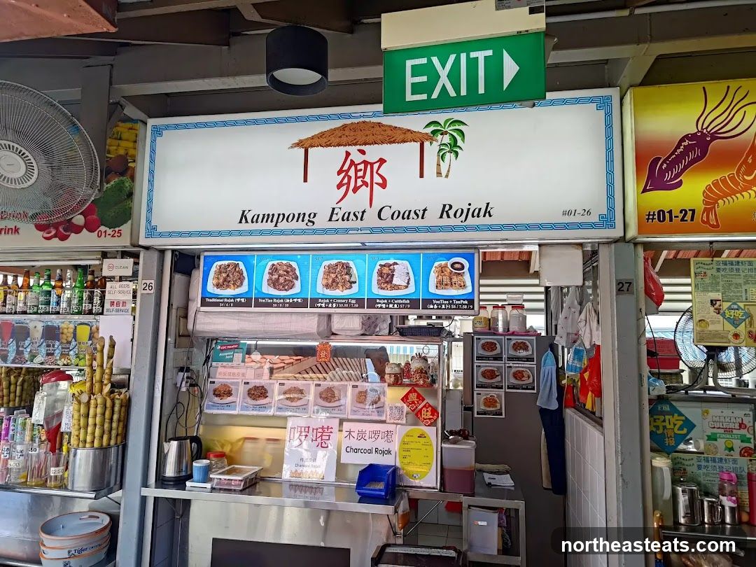A food stall with a large sign that reads "Kampong East Coast Rojak" and displays pictures of their dishes. Below the main sign, there are smaller signs with pictures and prices of various rojak options. To the left, there is a display of drinks and sugarcane. To the right, there are signs for other food stalls. An "EXIT" sign is visible above the main sign.