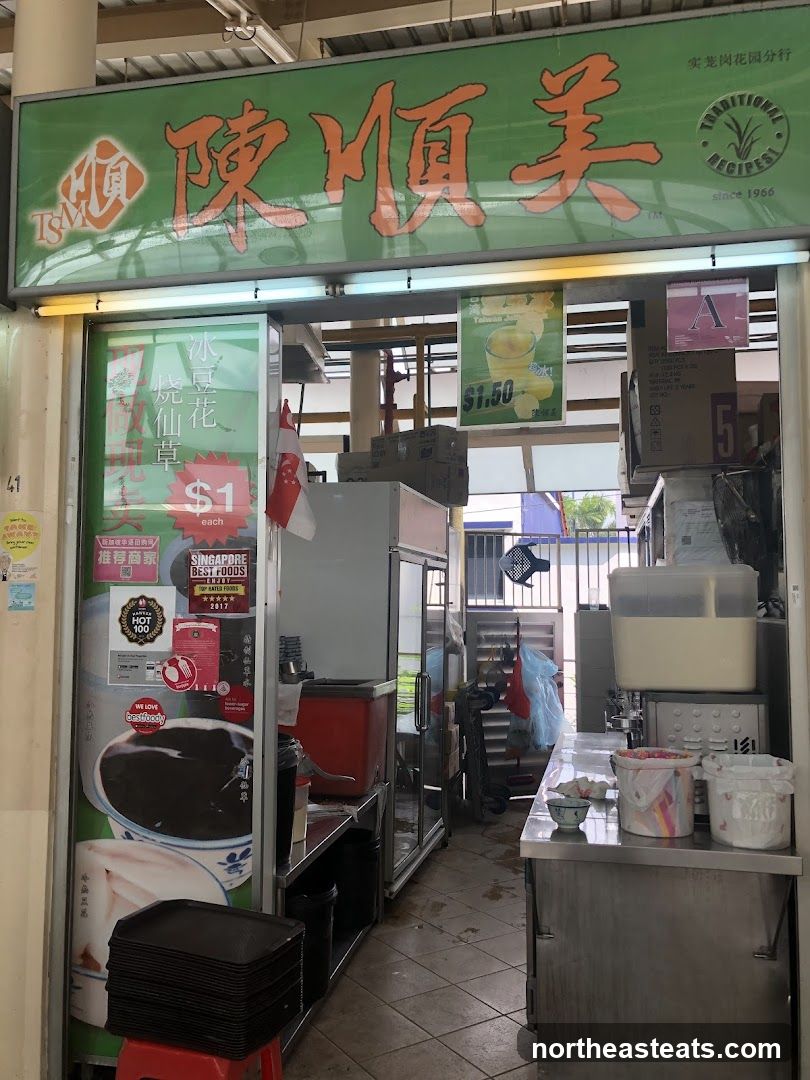 A dessert shop with a green awning and Chinese characters. There are posters and signs displayed on the glass door and inside the shop. A stack of black trays is visible on a red stool.