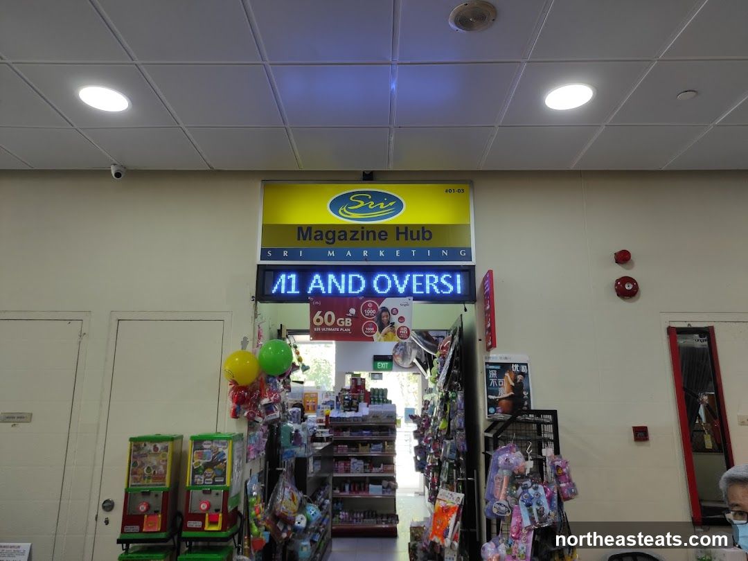 The image shows the entrance to a store called Sri Magazine Hub. Above the entrance, a yellow sign displays the store's name and logo. Below this, a digital sign flashes "11 AND OVERSI" and an advertisement for a 60GB data plan for $35. The store appears to sell magazines, snacks, and other convenience items.