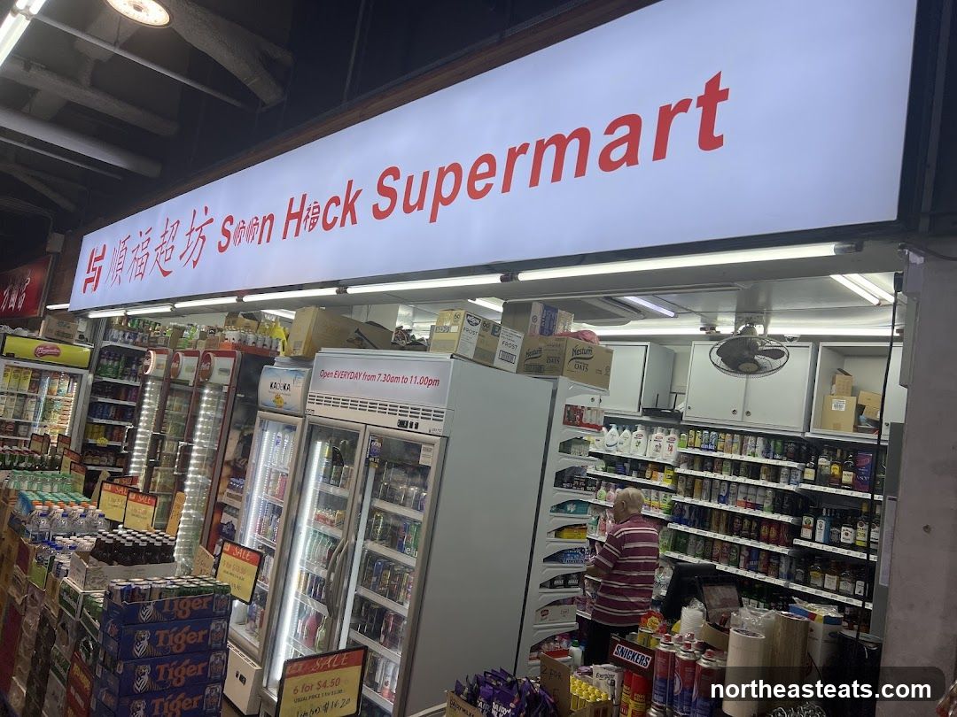 A brightly lit storefront for Soon Hack Supermart, with a large illuminated sign displaying the store name in red lettering. The entrance is open, revealing shelves stocked with various goods, including drinks in refrigerators, canned items, and packaged snacks. A person is visible inside, browsing the shelves. Signs indicate operating hours and sales promotions.