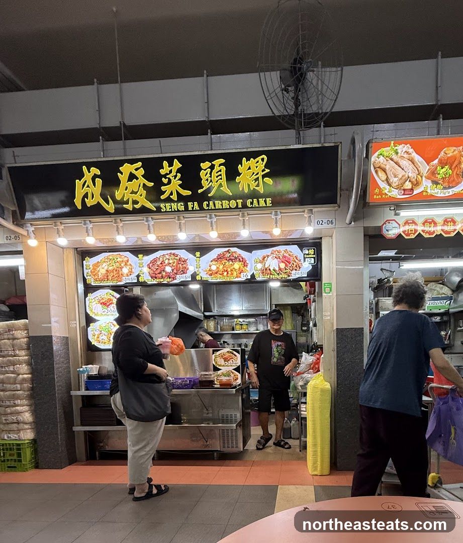 A food stall with a prominent sign reading "SENG FA CARROT CAKE" in English and Chinese characters. The stall displays images of various dishes, including carrot cake and other local delicacies, with prices listed. Several people are seen interacting with the stall, with some ordering food and others waiting. The stall appears to be located in an open-air food court or hawker center.