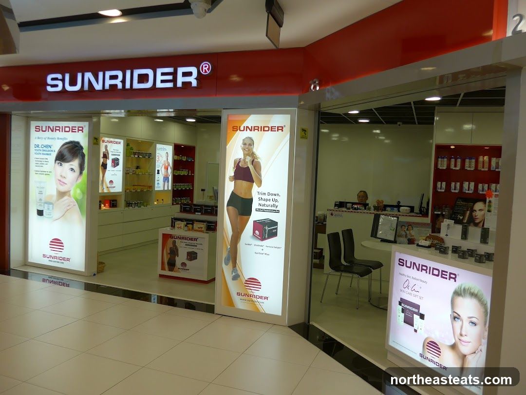 The exterior of a Sunrider store is shown, with illuminated displays featuring models and product information. The prominent "SUNRIDER" logo is visible above the entrance. Inside, shelves are stocked with various products.